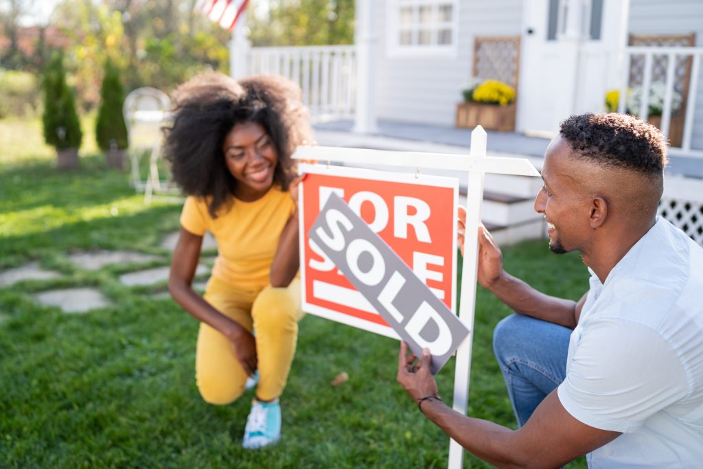 A black couple crouch outside a newly-purchased home. The female partner at the left, on one knee, watches, smiling, as the male partner on the right places a "sold" decal over the "for sale" sign in the lawn.
