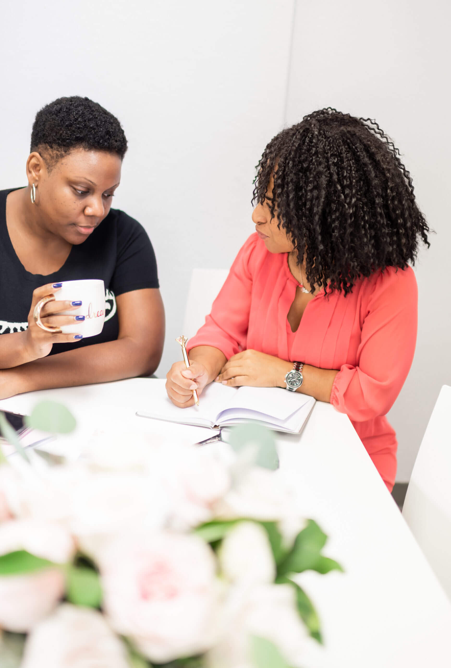 Woman, left, holding mug and looking down at book; Woman, right writing in book and looking back at other woman