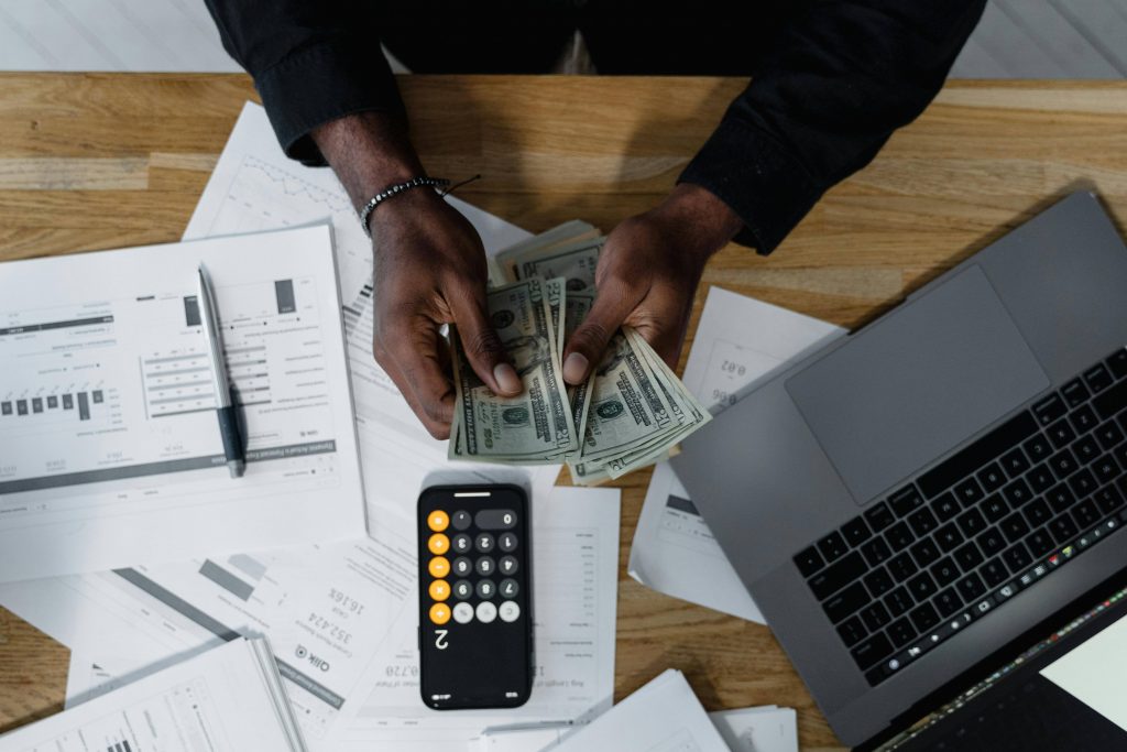 Shot of a person's hands counting bills, with a laptop open, papers, and a calculator in front of them